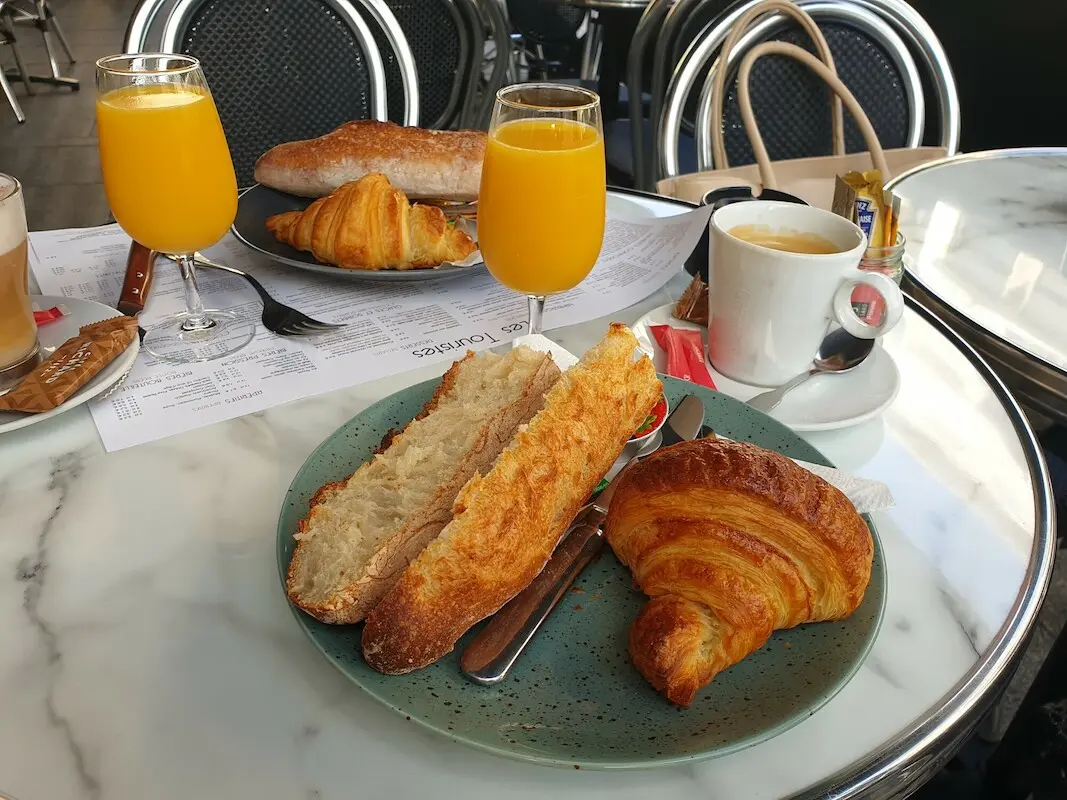 Frühstückstisch im Restaurant: Zwei Teller mit jeweils einem angerösteten Brötchen und einem Croissant und einem vollen Glas Orangensaft stehen auf einem Tisch. Dazu Kaffeee.