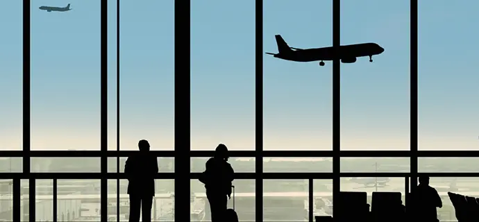 Großes Terminallfenster mit Ausblick auf das Vorfeld. Drei Personen-Silhouetten im Gegenlicht, beobachten startende Flugzeuge und das Geschehen am Himmel.