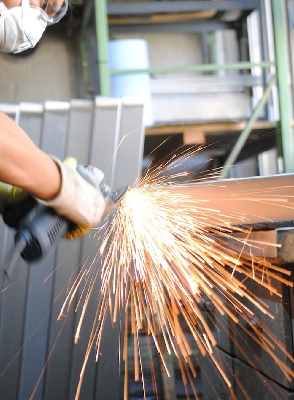 Ein Metallwerker schweißt gerade an einem Stück Metall. Funken fliegen, diese befinden sich in der Bildmitte und bieten das Haupt-Augenmerk.