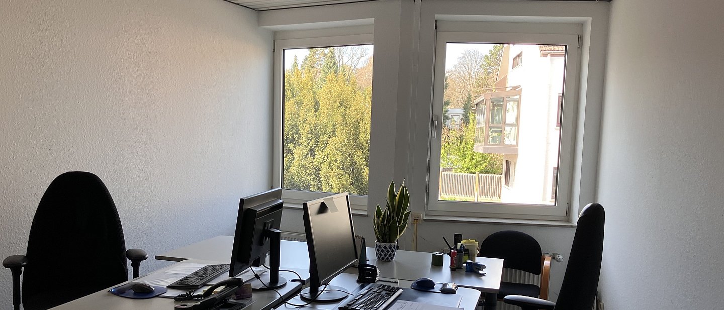 Zwei Schreibtische in einem Büro mit Computerbildschrimen und Schreibtischstühlen vor einem Fenster. Eine typisches Büro.