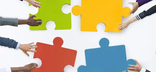 Mehrere Hände schieben bunte Puzzleteile zusammen. Die Szene symbolisiert Teamarbeit, Zusammenarbeit und das Zusammenfügen unterschiedlicher Menschen oder Ideen.