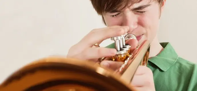 Ein Jugendlicher spielt konzentriert Trompete. Sein Gesicht ist angespannt, die Augen geschlossen, während er die Ventile des Instruments drückt. Die goldene Trompete ist im Vordergrund prominent sichtbar.