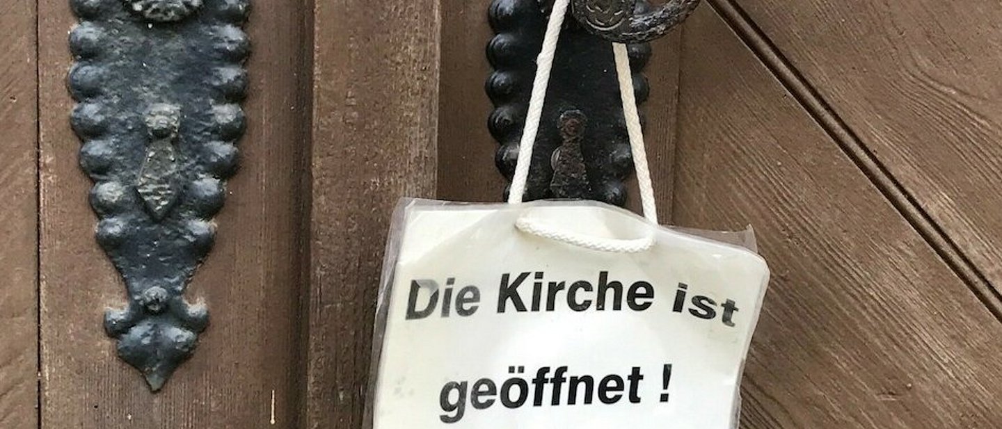 Weißes Schild hängt an Klinke der Kirchentür. Auf dem Schild steht: "Die Kirche ist geöffnet".