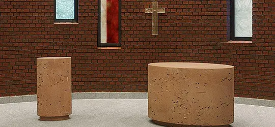 Altarraum mit steinernem Altar und Taufstein vor einer Backsteinwand mit Kreuz.