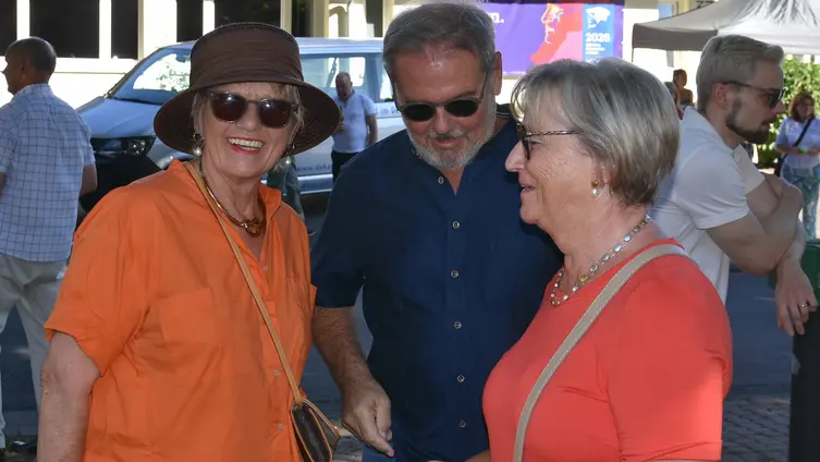 Zwei Frauen, beide orange gekleidet, in ihrer Mitte ein Mann, unterhalten sich lachend. Die linke Frau trägt Hut und Sonnenbrille und lacht in die Kamera.