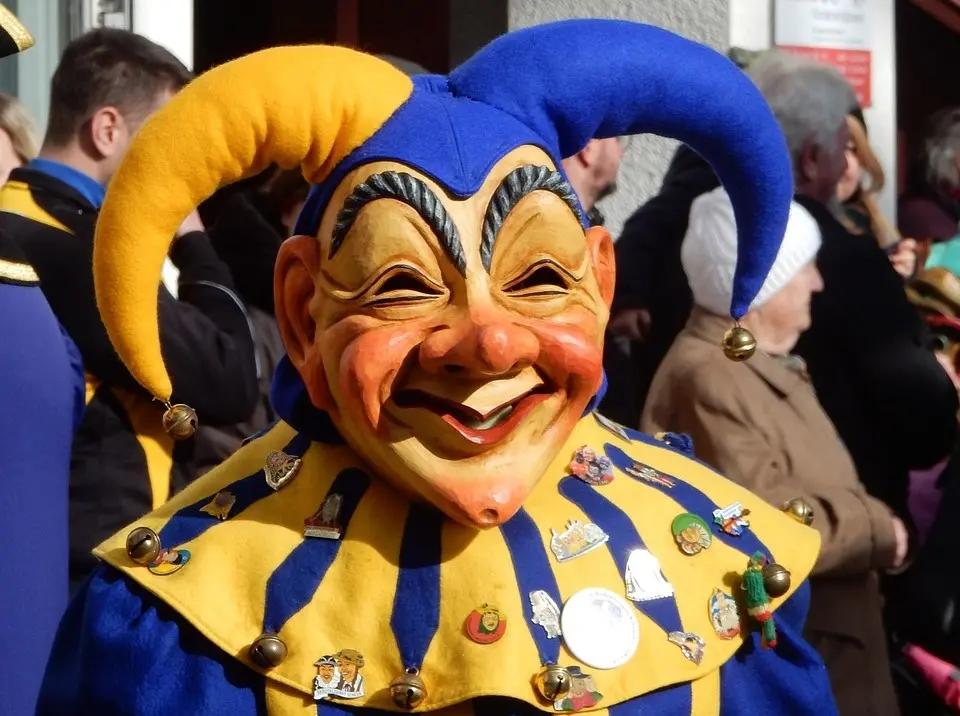 Ein Narr mit einer großen, freundlichen Maske und einem Hut mit großer gelber und blauer Bommel.