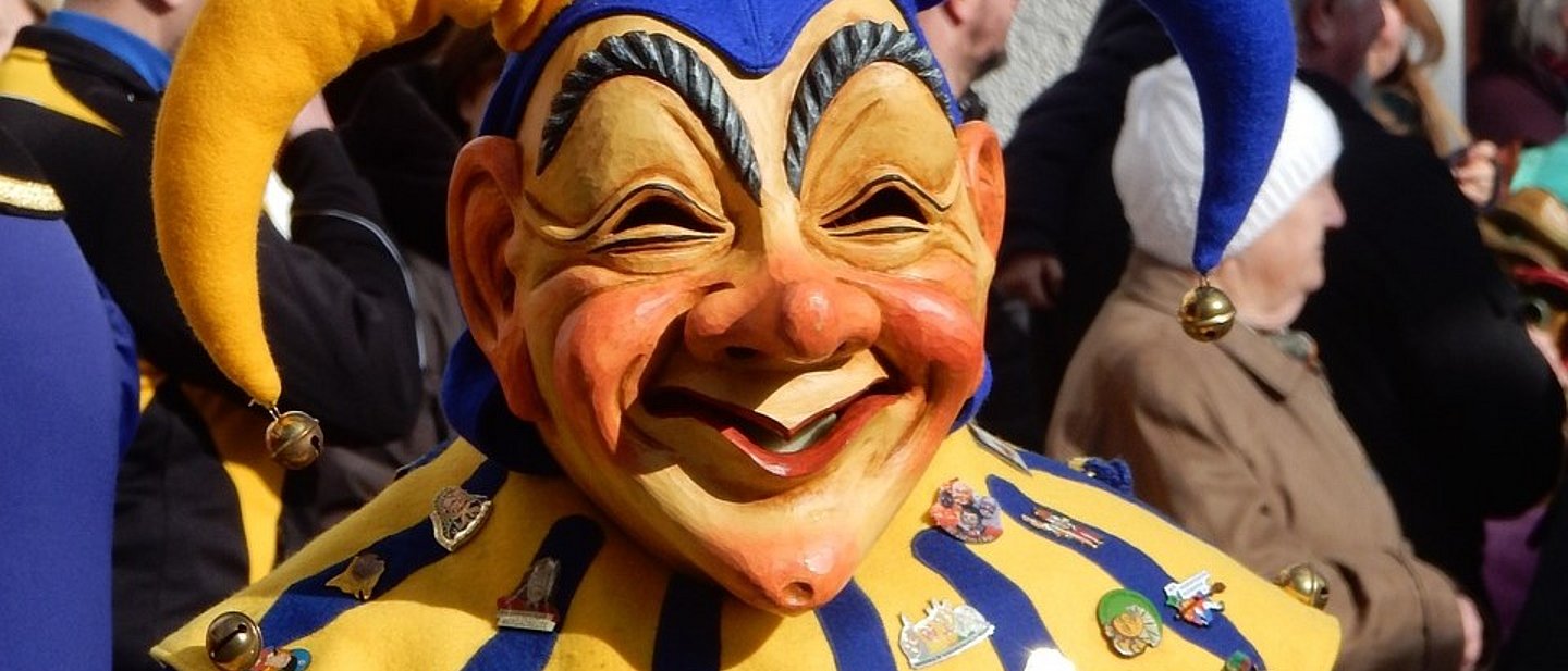Ein Narr mit einer großen, freundlichen Maske und einem Hut mit großer gelber und blauer Bommel.