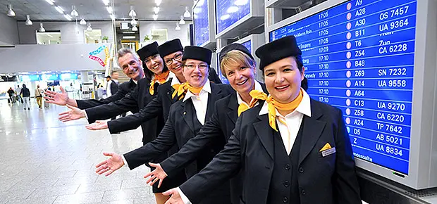 Mehrere Mitarbeitende in Uniform stehen lächelnd in einer Reihe vor einer großen Anzeigetafel am Flughafen und begrüßen mit ausgestreckten Armen die ankommenden Reisenden. Die Szene vermittelt eine freundliche und einladende Atmosphäre im Terminal.