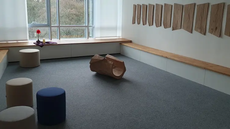 Das Foto zeigt die Totale des Raums, mit dem Holzstamm in der Mitte, Sitzgelegenheiten und den Holzscheiben an der Wand.