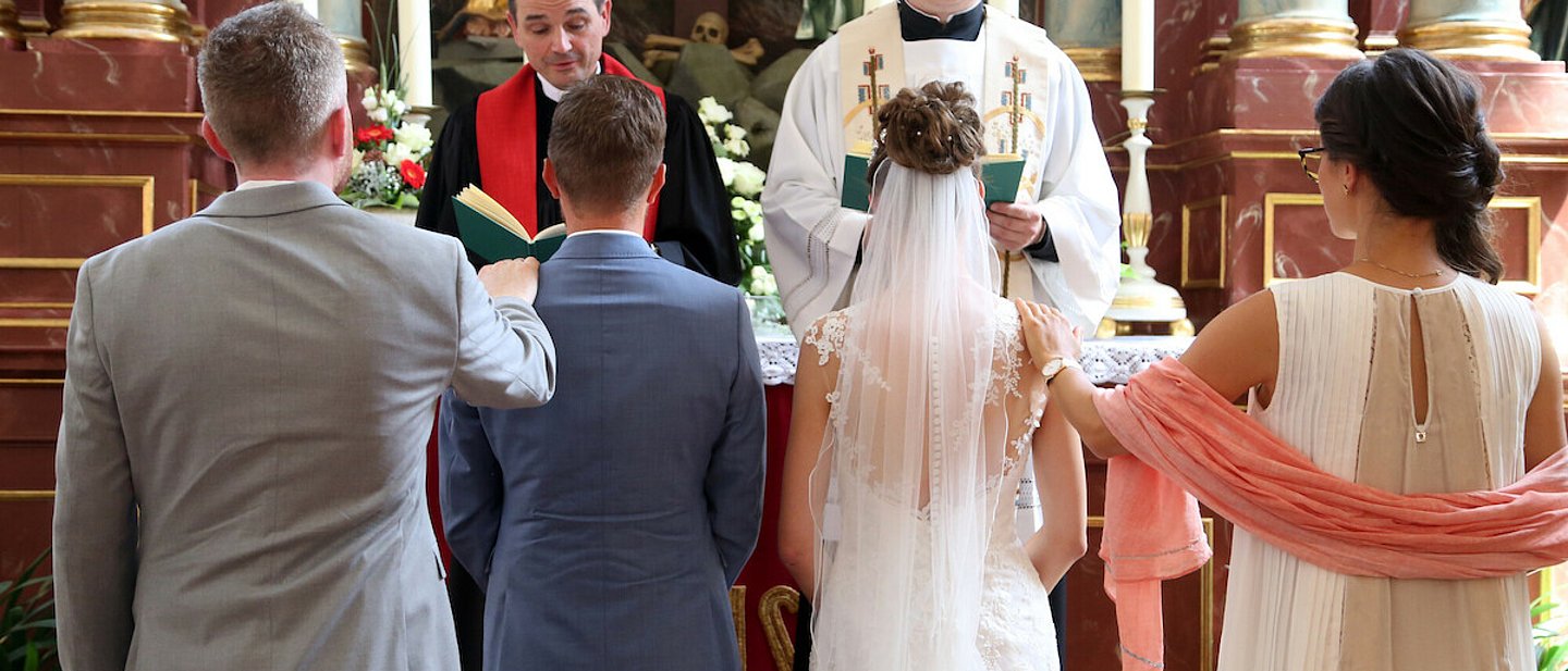 Ehepaar und Trauzeugen stehen im Altarraum. Vor ihnen katholischer und evangelischer Pfarrer. Im Hintergrund der Hochaltar mit Kerzen