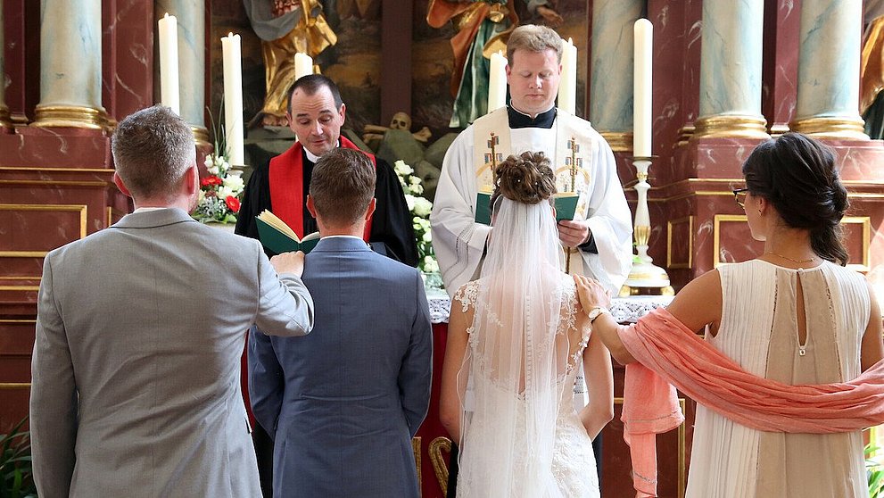 Ehepaar vor Altar Ehepaar und Trauzeugen stehen im Altarraum. Vor ihnen katholischer und evangelischer Pfarrer. Im Hintergrund der Hochaltar mit Kerzen