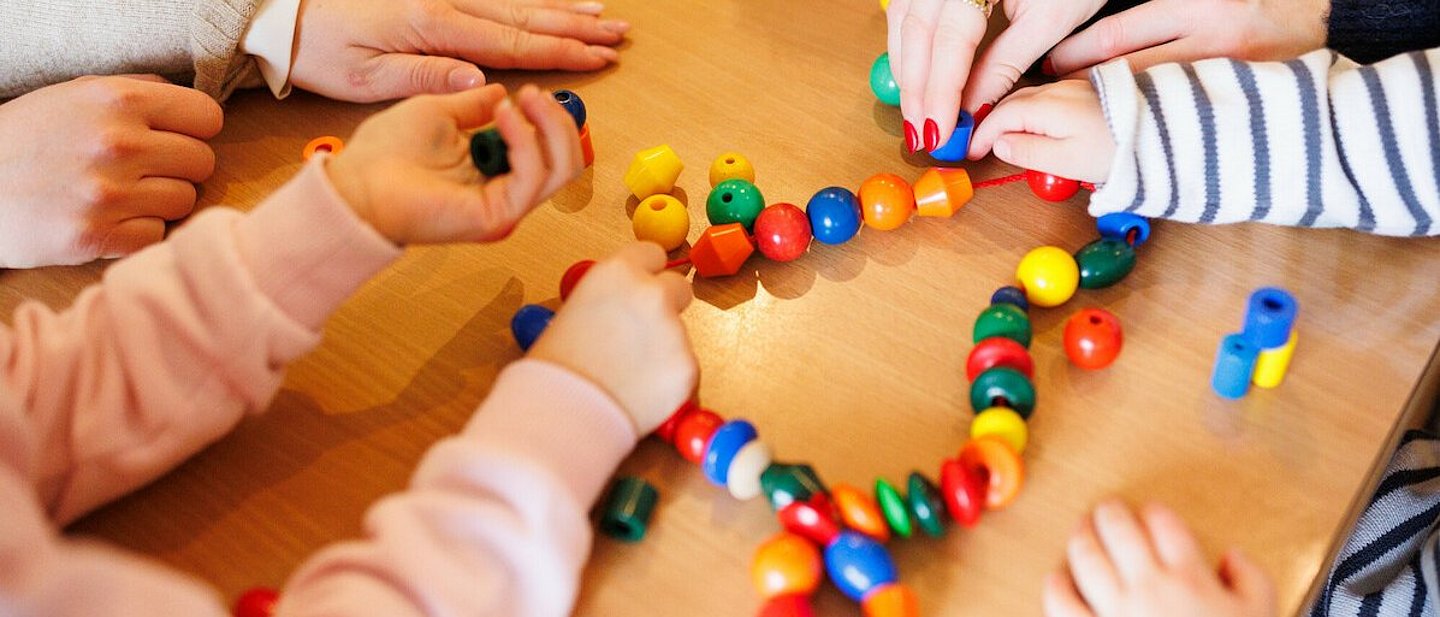 Kita - Kinder beim Spielen. An einem Tisch sitzen einige Kinder, eine Erzieherin und spielen mit einer Kette mit bunten Kugeln und anderen Formen, die auf einer Schnur aufgereiht sind.