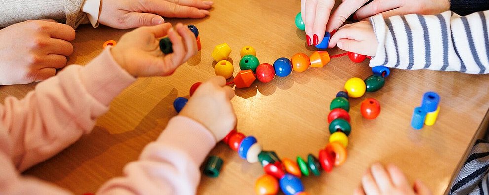 Kita - Kinder beim Spielen. An einem Tisch sitzen einige Kinder, eine Erzieherin und spielen mit einer Kette mit bunten Kugeln und anderen Formen, die auf einer Schnur aufgereiht sind.