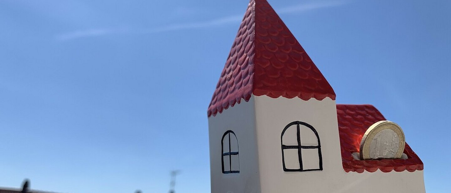 Spardose in Form einer weißen Kirche mit rotem Dach. Eine 1 € -Münze wird gerade durch den Schlitz geschoben