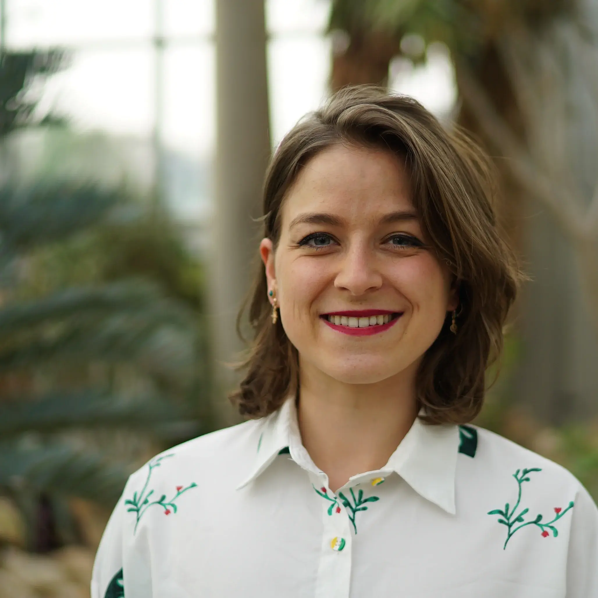 Ein Portrait von Alexandra Hild in weißer Bluse vor einem unscharfen Hintergrund mit Palmen in einem Gewächshaus