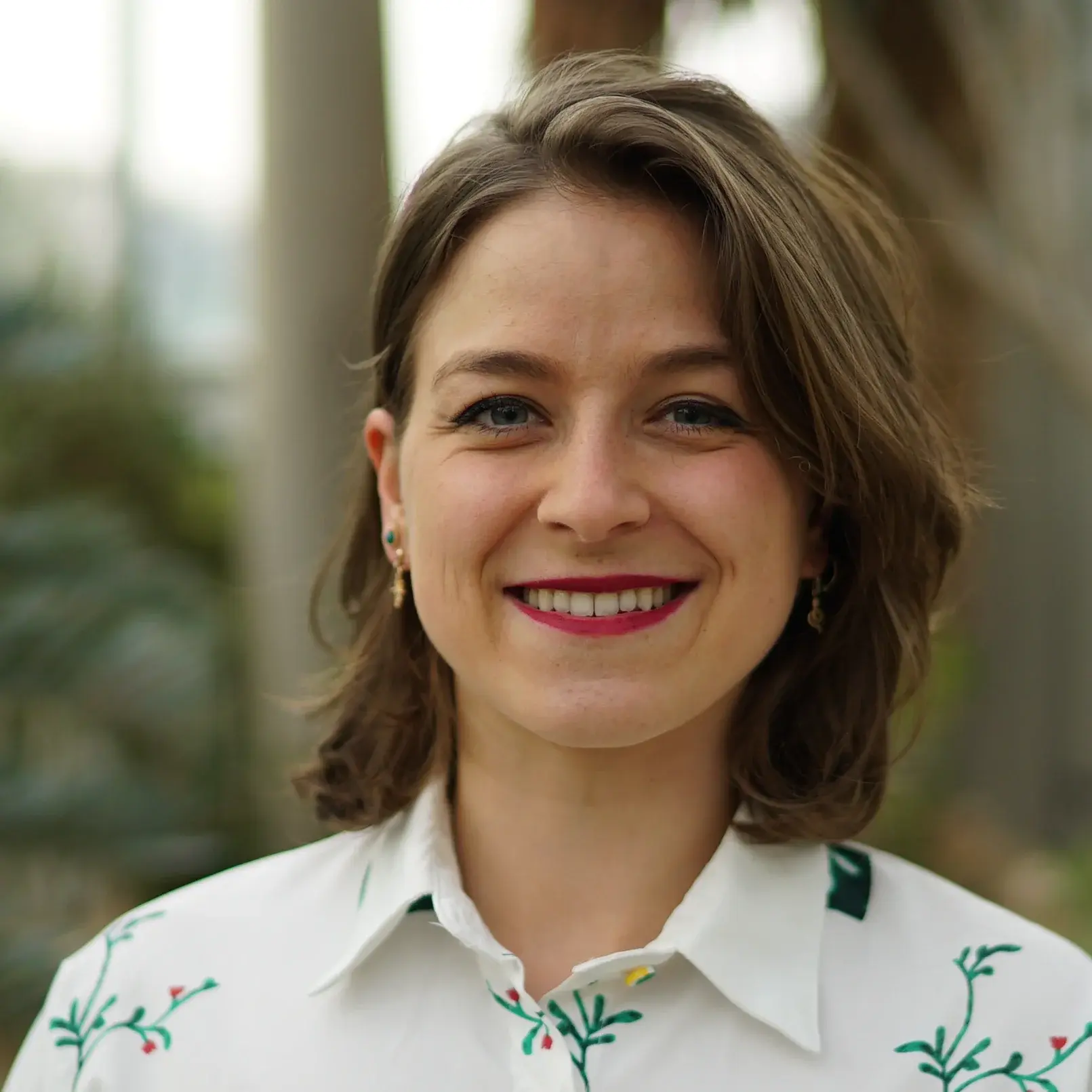 Ein Portrait von Alexandra Hild in weißer Bluse vor einem unscharfen Hintergrund mit Palmen in einem Gewächshaus