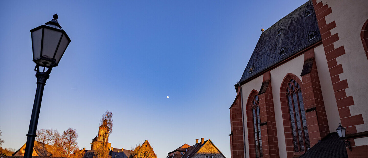 Man sieht einen Teil der Marienkirche in Büdingen auf der rechten Seite des Bildes. Auf der linken Seite steht eine Straßenlaterne im historischen Design. dazwischen ist viel Freiraum und blauer Himmel. Im Hintergrund sieht man mehrere historische Gebäude.