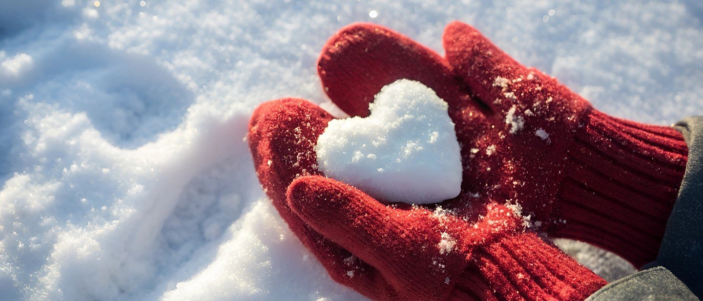 2 Hände in roten Wollfäustlingen halten ein aus Schnee geformtes Herz