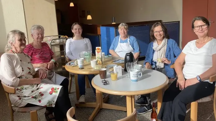 Das Bild zeigt eine Gruppe von sechs Frauen, die um zwei runde Tische sitzen. Auf den Tischen stehen Tassen, Krüge und eine Packung Saft, was darauf hindeutet, dass sie zusammen Kaffee trinken oder eine Mahlzeit genießen. Die Frauen sind unterschiedlich gekleidet, einige tragen Alltagskleidung, während eine eine Schürze trägt, was möglicherweise darauf hindeutet, dass sie die Gastgeberin ist oder an der Zubereitung beteiligt war. Sie scheinen entspannt und engagiert in Gesprächen zu sein. Der Raum wirkt hell und einladend mit natürlichem Licht.