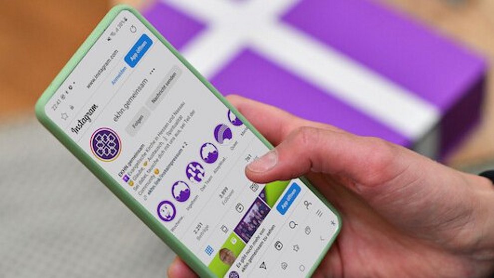 Pfarrerin Nele Schomakers hält ein Smartphone in ihrer Hand. Im unscharfen Hintergrund ist ein weißes Kreuz auf einem lila Untergrund zu erkennen.