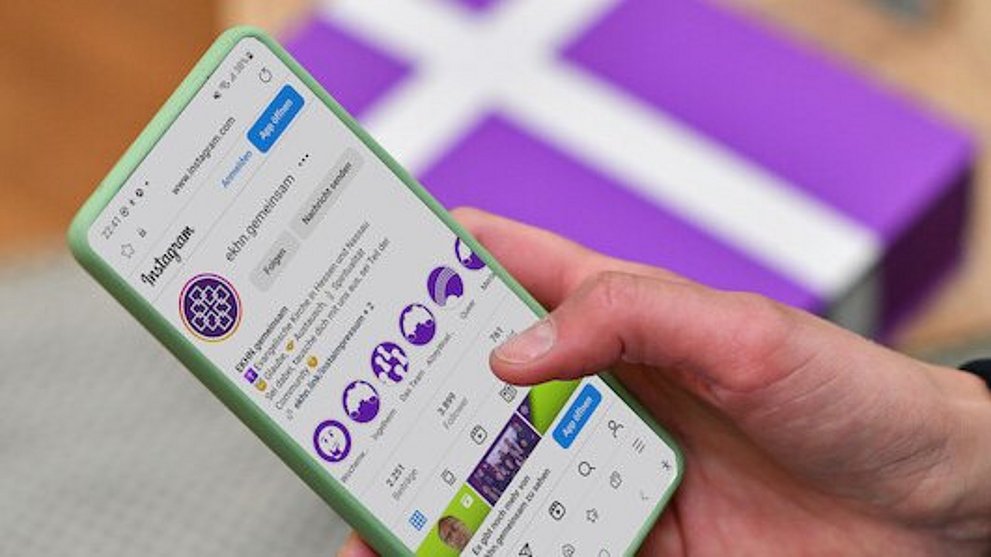 Pfarrerin Nele Schomakers hält ein Smartphone in ihrer Hand. Im unscharfen Hintergrund ist ein weißes Kreuz auf einem lila Untergrund zu erkennen.