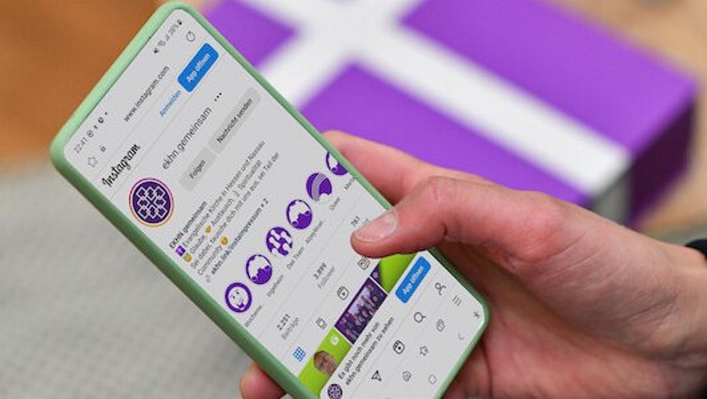 Pfarrerin Nele Schomakers hält ein Smartphone in ihrer Hand. Im unscharfen Hintergrund ist ein weißes Kreuz auf einem lila Untergrund zu erkennen.