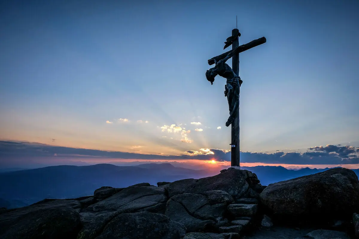 Man sieht einen Berggipfel mit dem dem überlebensgroßen Christus am Kreuz vor einem Abendhimmel mit Sonnenuntergang