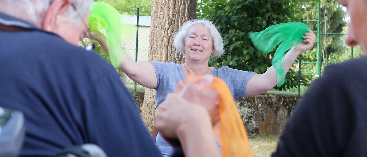 Dorothee Ewald steht strahlend vor Senior*innen und hält zwei grüne Tücher in die Höhe.