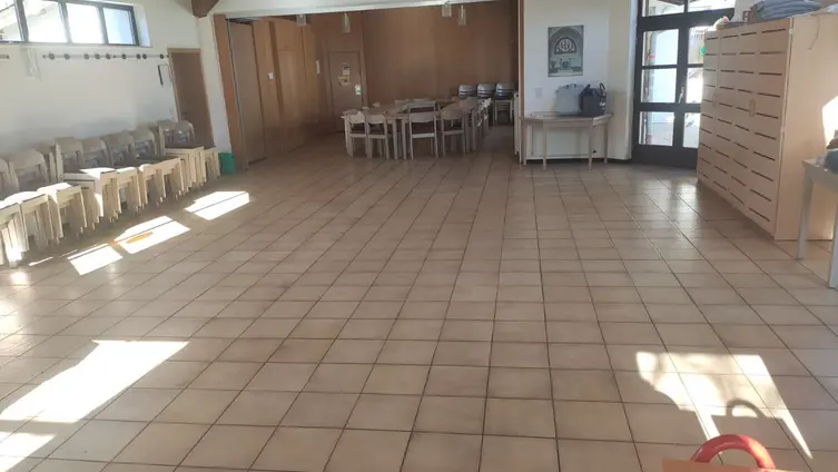 Ein großer, gefließter Raum. Im Hintergrund steht eine Tischgruppe mit Stühlen.