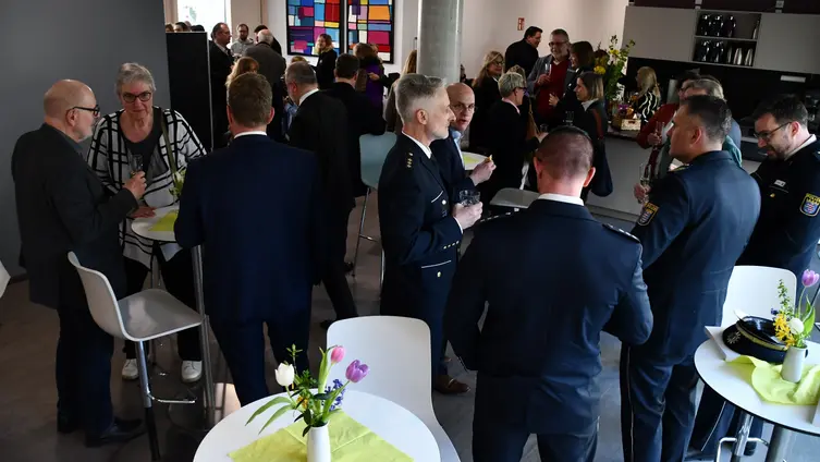 Viele Menschen, einige in Polizeiuniform, stehen an Stehtischen und reden miteinander. 