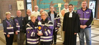 Gruppenfoto in einer Kirche: Mehrere Personen in lila Einsatzjacken stehen mit Geistlichen zusammen. Zwei Frauen halten eine große Kerze mit einem bunten Symbol. Im Hintergrund ist ein Altar mit Kreuz zu sehen.