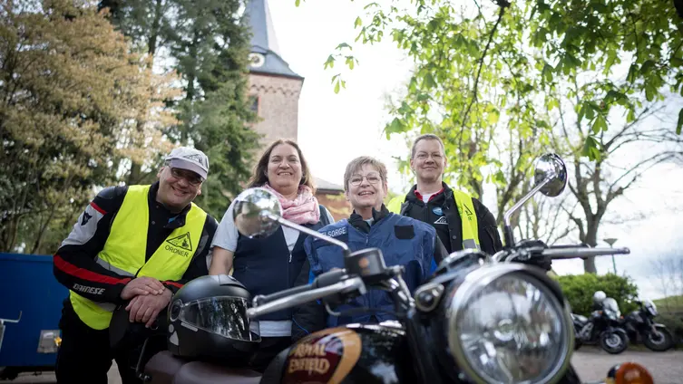 Die vier Personen stehen hinter einem Motorrad. Im Hintergrund ist die Kirche zu sehen.