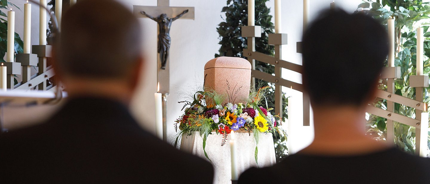 Das Foto zeigt ein schwarz gekleidetse PAaar von hinten. Es sitzt in einer Trauerhalle vor einer Urne und Blumen. An der Wand dahinter hängt ein Kreuz mit Jesus.