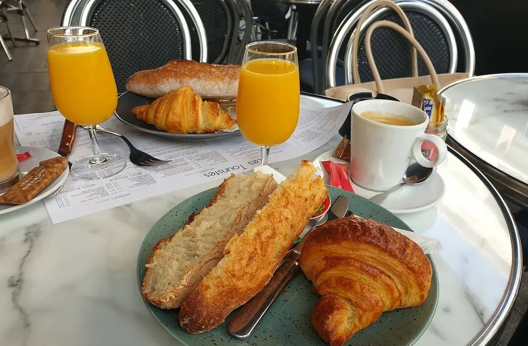 Frühstückstisch im Restaurant: Zwei Teller mit jeweils einem angerösteten Brötchen und einem Croissant und einem vollen Glas Orangensaft stehen auf einem Tisch. Dazu Kaffeee.