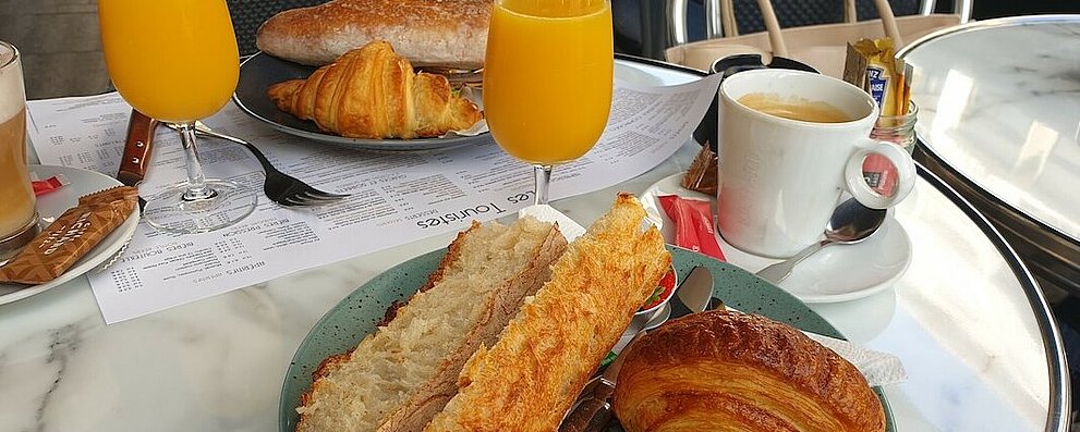 Frühstückstisch im Restaurant: Zwei Teller mit jeweils einem angerösteten Brötchen und einem Croissant und einem vollen Glas Orangensaft stehen auf einem Tisch. Dazu Kaffeee.