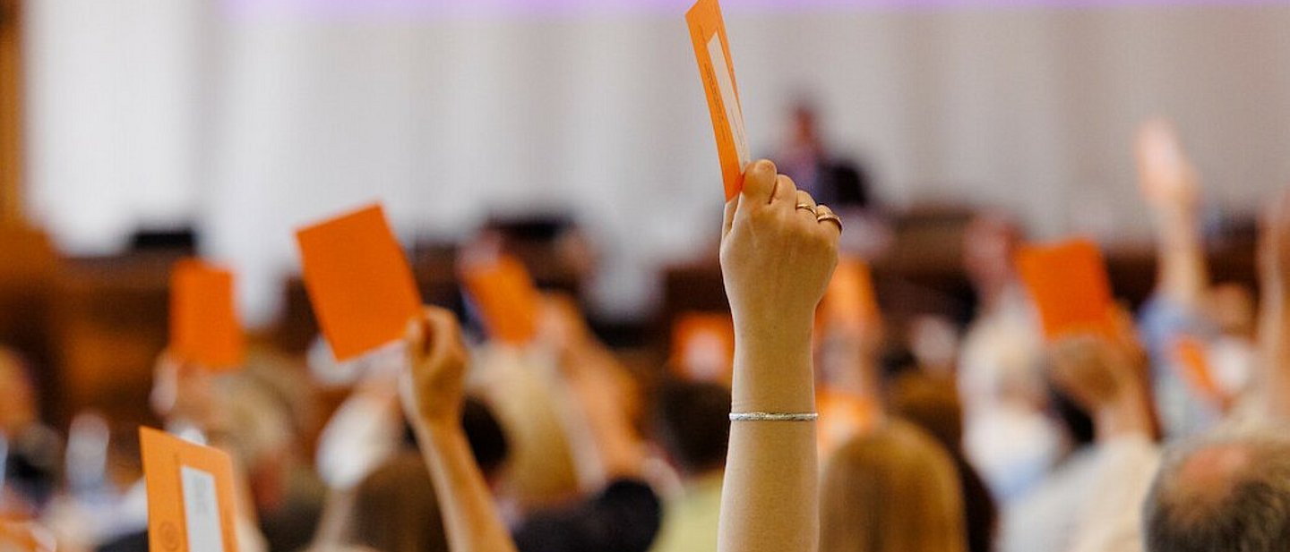Man sieht auf Hände, die einen orangenen Abstimmzettel halten