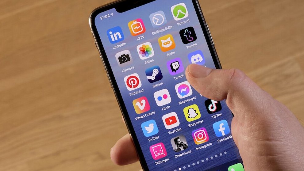 Man sieht ein Handy/IPhone in einer Hand mit den verschiedensten Apps für Socialmedia vor einem Holzhintergrund.
