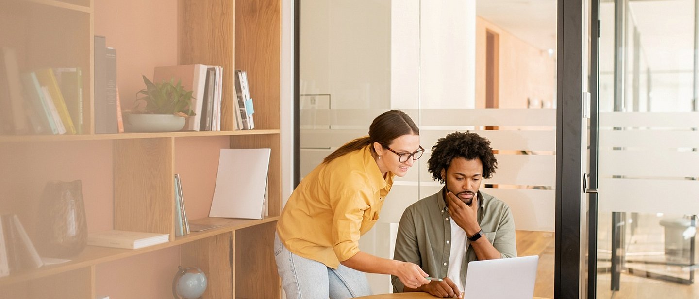 Frau mit dunklen Haaren und gelber Bluse erklärt Mann mit dunklem Haaren etwas am Laptop 