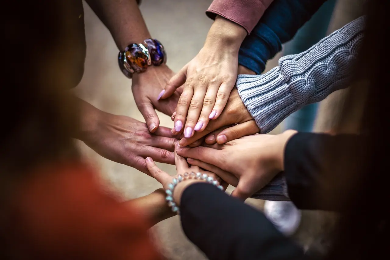 Das Bild zeigt eine Gruppe von Händen, die in der Mitte aufeinander liegen. Die Hände gehören verschiedenen Personen, die eng beieinander sitzen. Die Hände sind vielfältig und zeigen unterschiedliche Hautfarben und Schmuckstücke. Das Bild suggeriert Einheit, Zusammenarbeit und Gemeinschaft.