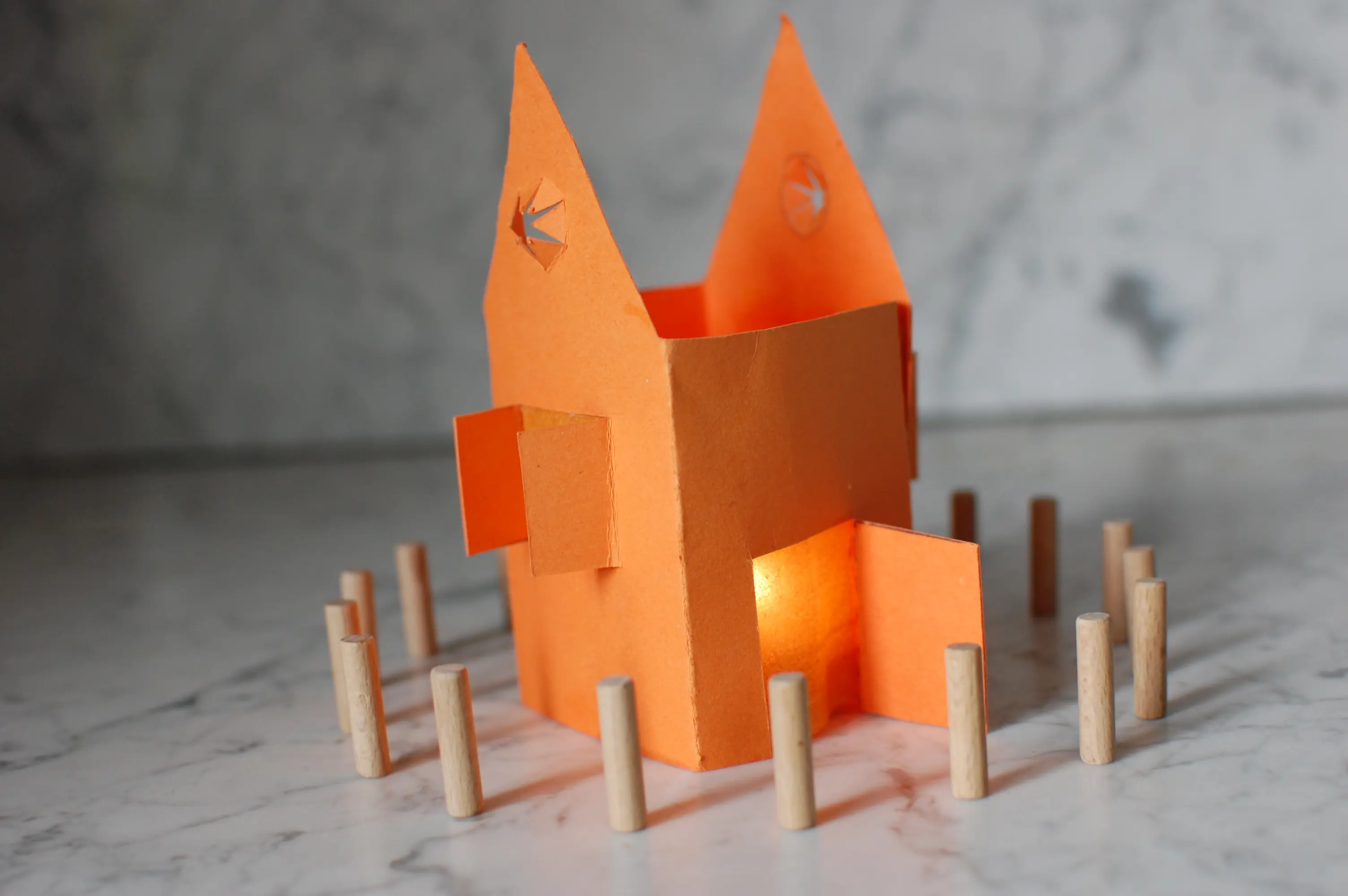 Das Foto zeigt Holzstäbchen die im Kreis um ein leuchtendes Papierhaus stehen.