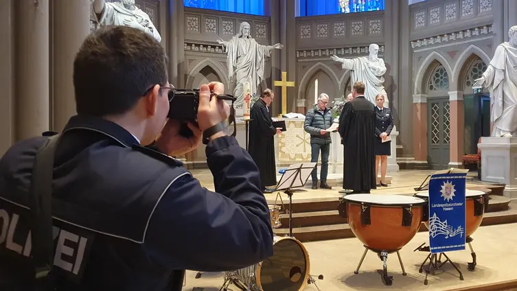 Links ist ein Polizist in Uniform von hinten zu sehen, er fotografiert. Rechts im Hintergrund der Altarraum, wo der Pfarrer von mehreren Personen gesegnet wird.