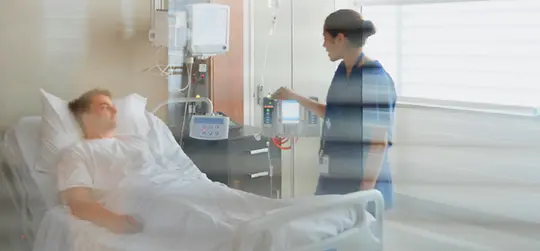 Pflegekraft steht am Bett eines Patienten in einem Krankenhauszimmer und bedient ein medizinisches Gerät.