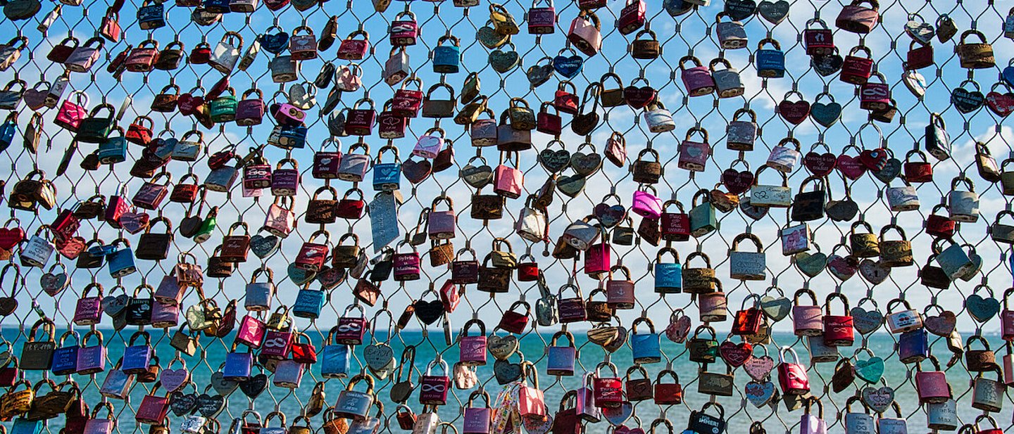 Viele bunte Liebesschlösser mit einigen Gravuren hängen in einem Netz. Im Hintergrund ist das Meer zu sehen.