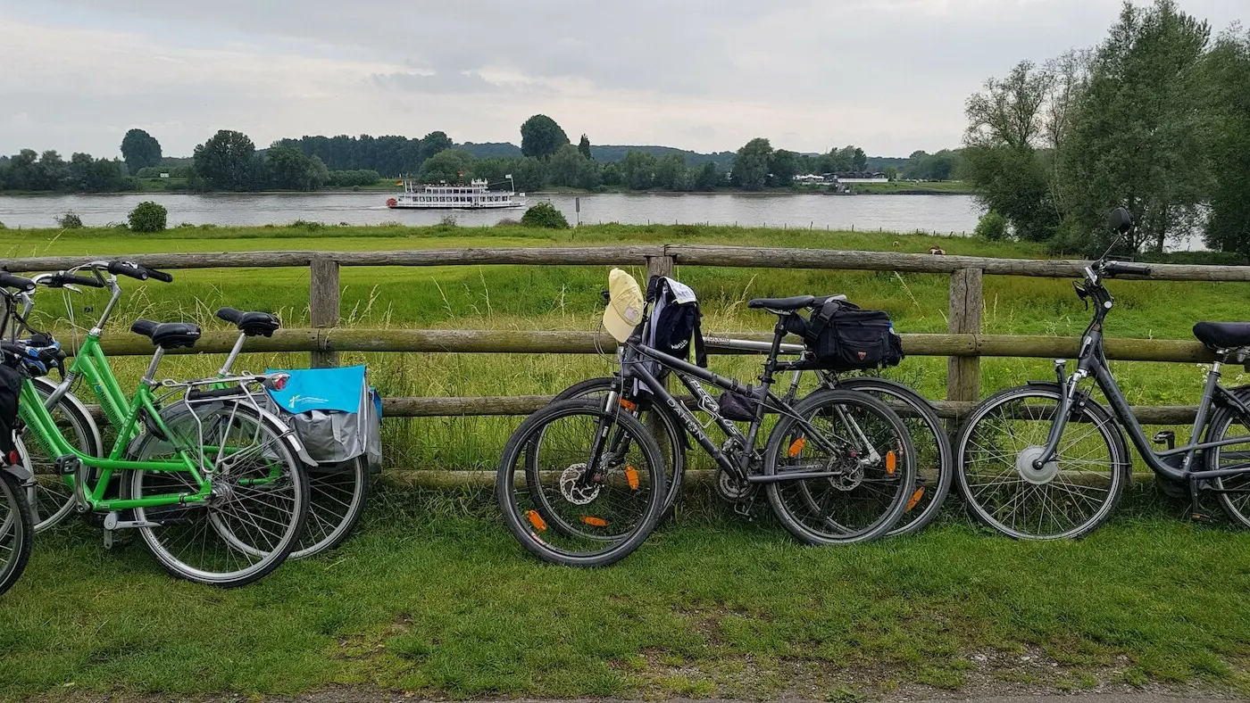 Fünf Fahrräder stehen an einen Holzzaun angelehnt. Hinter dem Zaun ist eine Weide zu sehen, dahinter ein Fluss mti einem Boot.