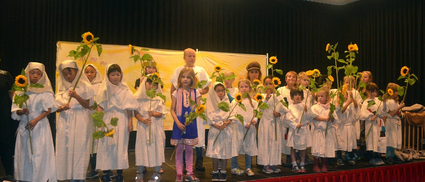 Bild zeigt einen Kinderchor auf der Bühne, in weißen Kostümkitteln und Sonnenblumen