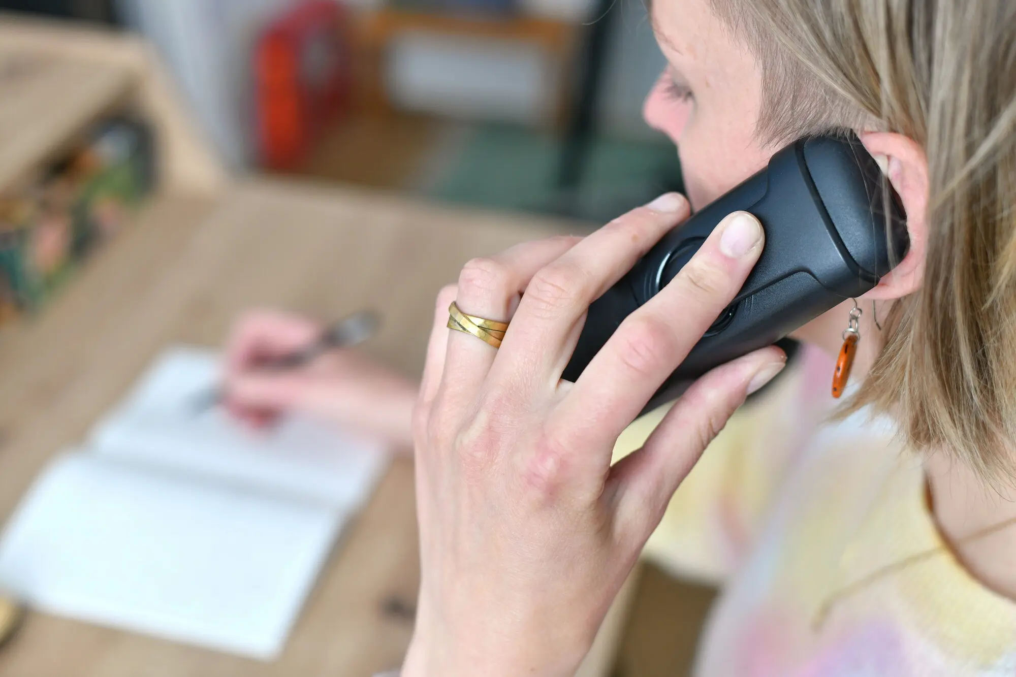 Eine Person hält einen Telefonhörer  nah an das Ohr, während sie zuhört. Im Hintergrund ist eine Hand mit einem Stift zu sehen, die Notizen auf einen Notizblock schreibt. Die Szene wirkt wie ein konzentriertes Telefongespräch.