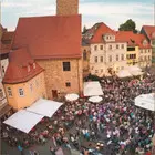 Luftaufnahme eines belebten Platzes in einer Altstadt mit vielen Menschen, Sonnenschirmen und historischen Gebäuden.