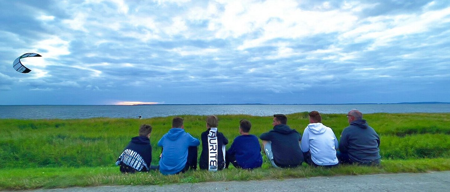 Man sieht eine Gruppe Jugendlicher am Strand sitzen die einem Kiter bei Ihrem Sport zusehen. Im Hintergrund sieht man den mit Gras bewachsenen Strand mit dem Meer dahinter. Der Drachen des Kiters fliegt in dem bewölktem Himmel auf der linken Seite des Bildes.