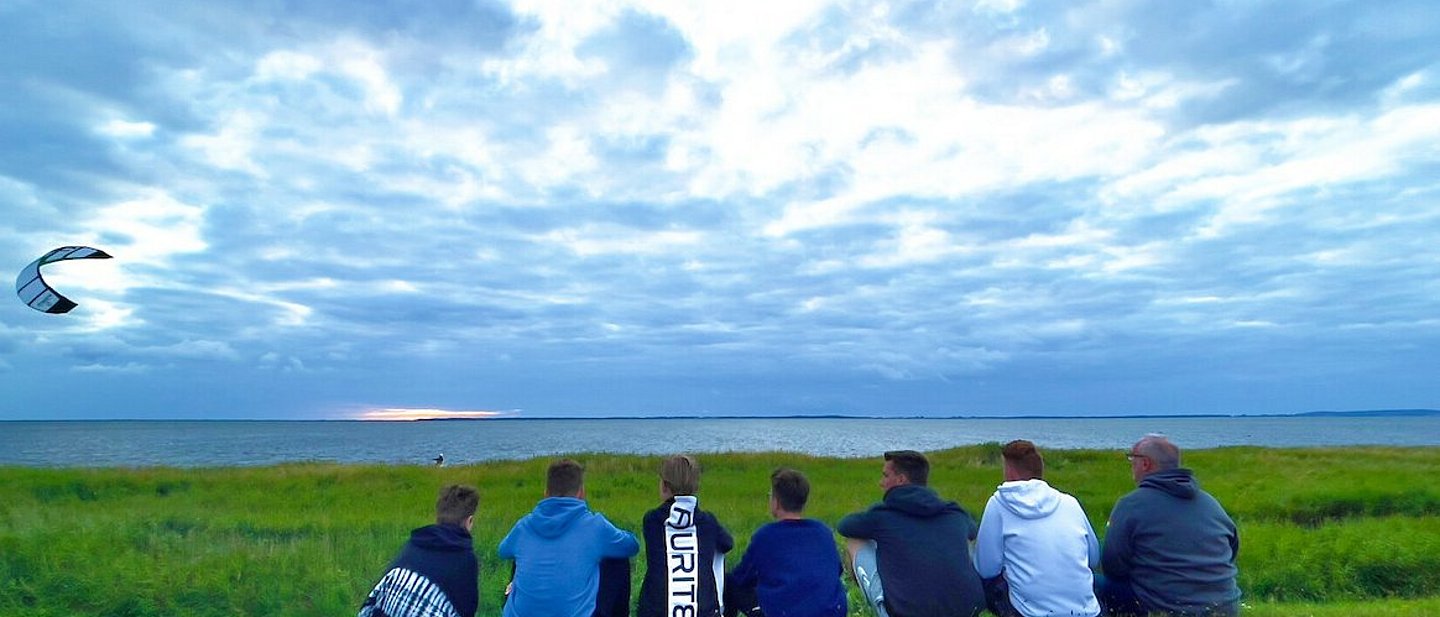Man sieht eine Gruppe Jugendlicher am Strand sitzen die einem Kiter bei Ihrem Sport zusehen. Im Hintergrund sieht man den mit Gras bewachsenen Strand mit dem Meer dahinter. Der Drachen des Kiters fliegt in dem bewölktem Himmel auf der linken Seite des Bildes.