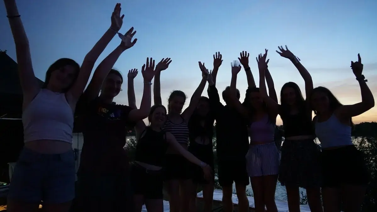 Man sieht eine Gruppe Jugendliche vor dem Abendhimmel, die Gesichter verschattet, die die Arme hoch reißen